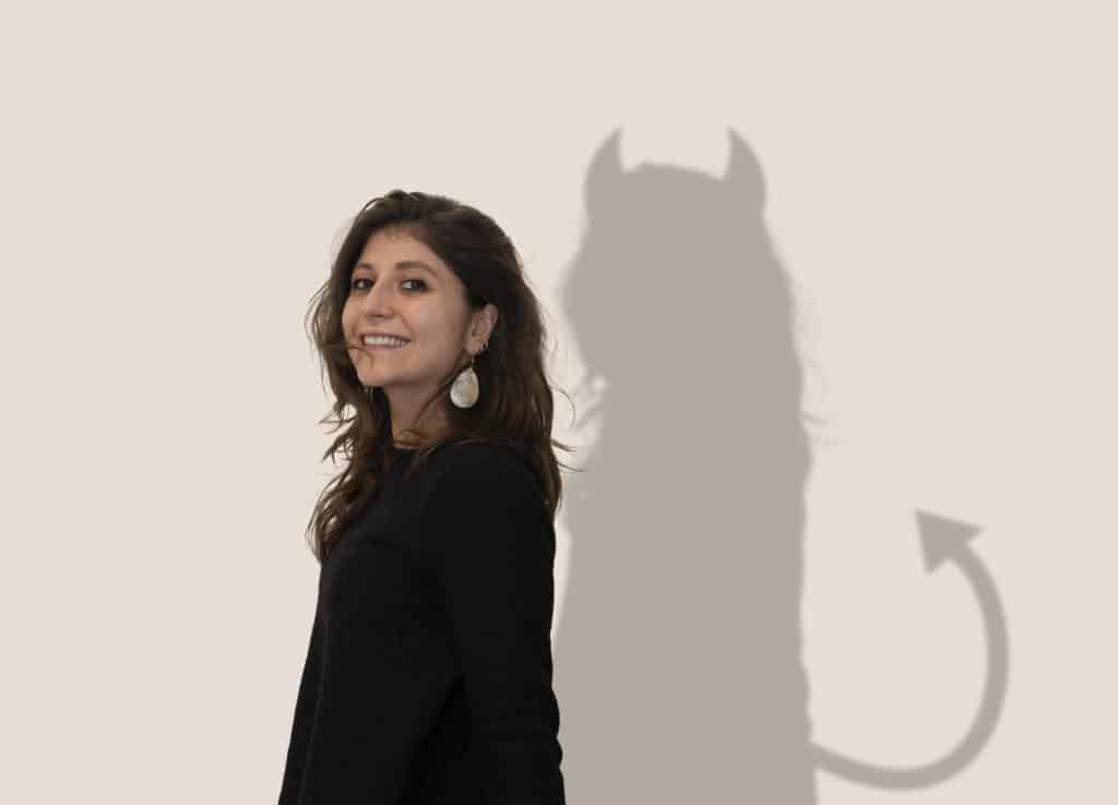 Smiling woman against a wall. Her shadow has horns and a pointy tail.
